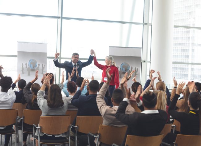Rear view of diverse business people applauding and celebrating while they are sitting in front of mixed race and Caucasian business people at business seminar in office building