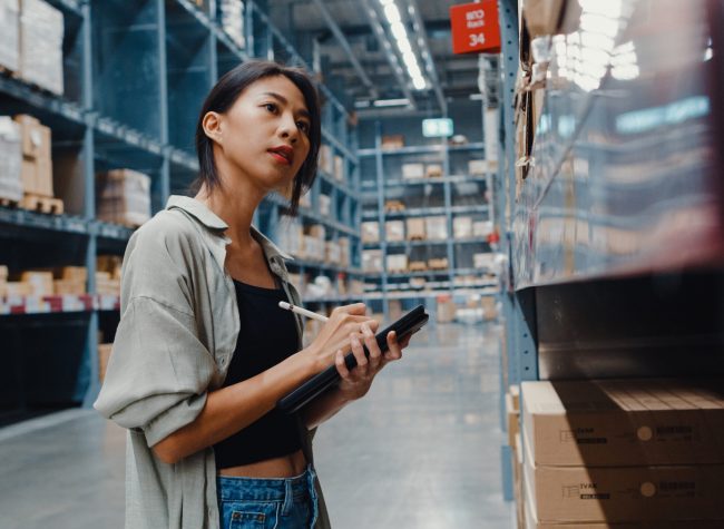 Attractive young Asia businesswoman manager looking for goods using digital tablet checking inventory levels standing in retail shopping center. Distribution, Logistics, Packages ready for shipment.